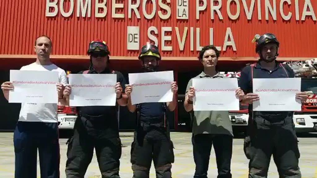 Cinco bomberos posan delante de la puerta del hangar con un papel en las manos cada uno que pone: "Condenados a salvar vidas".