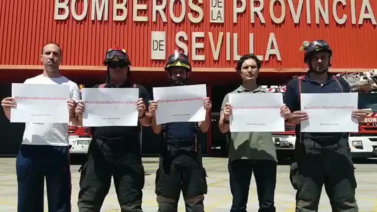 Cinco bomberos posan delante de la puerta del hangar con un papel en las manos cada uno que pone: "Condenados a salvar vidas".