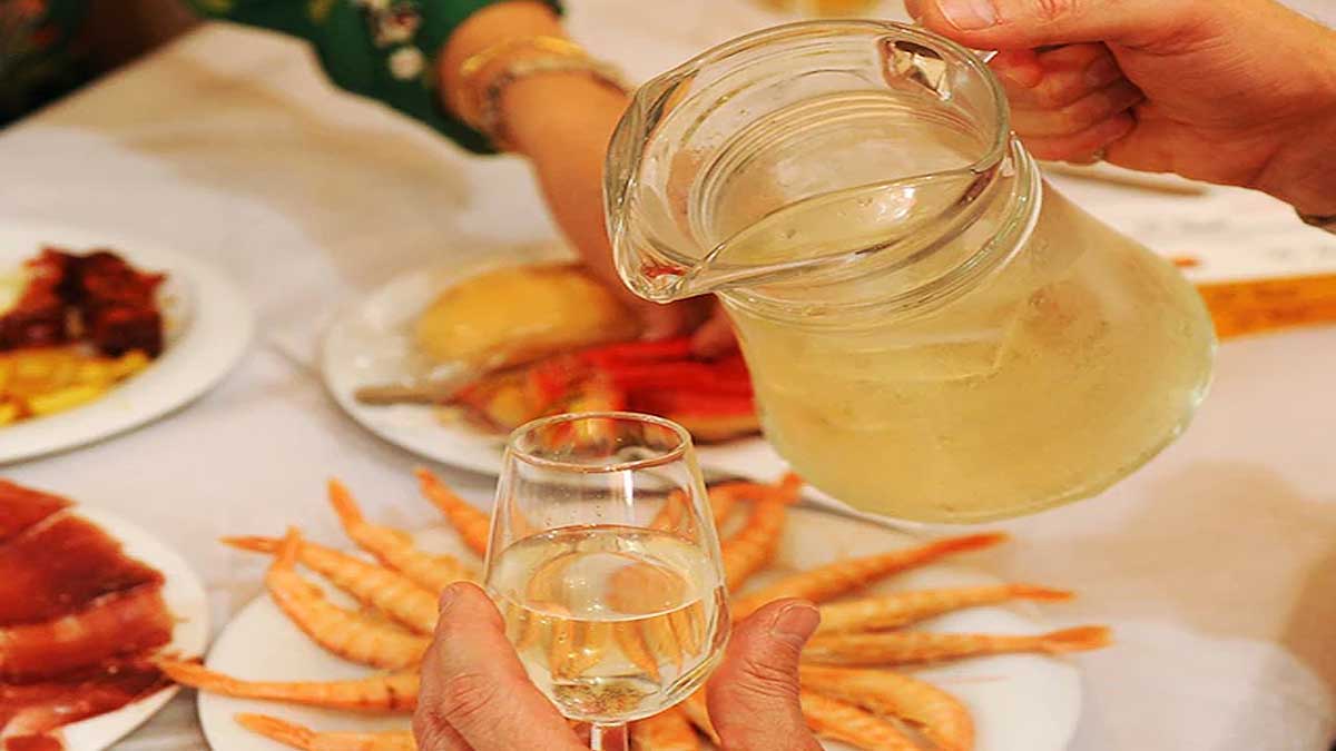 Hombre sirviendo una copa de rebujito con comida al fondo.