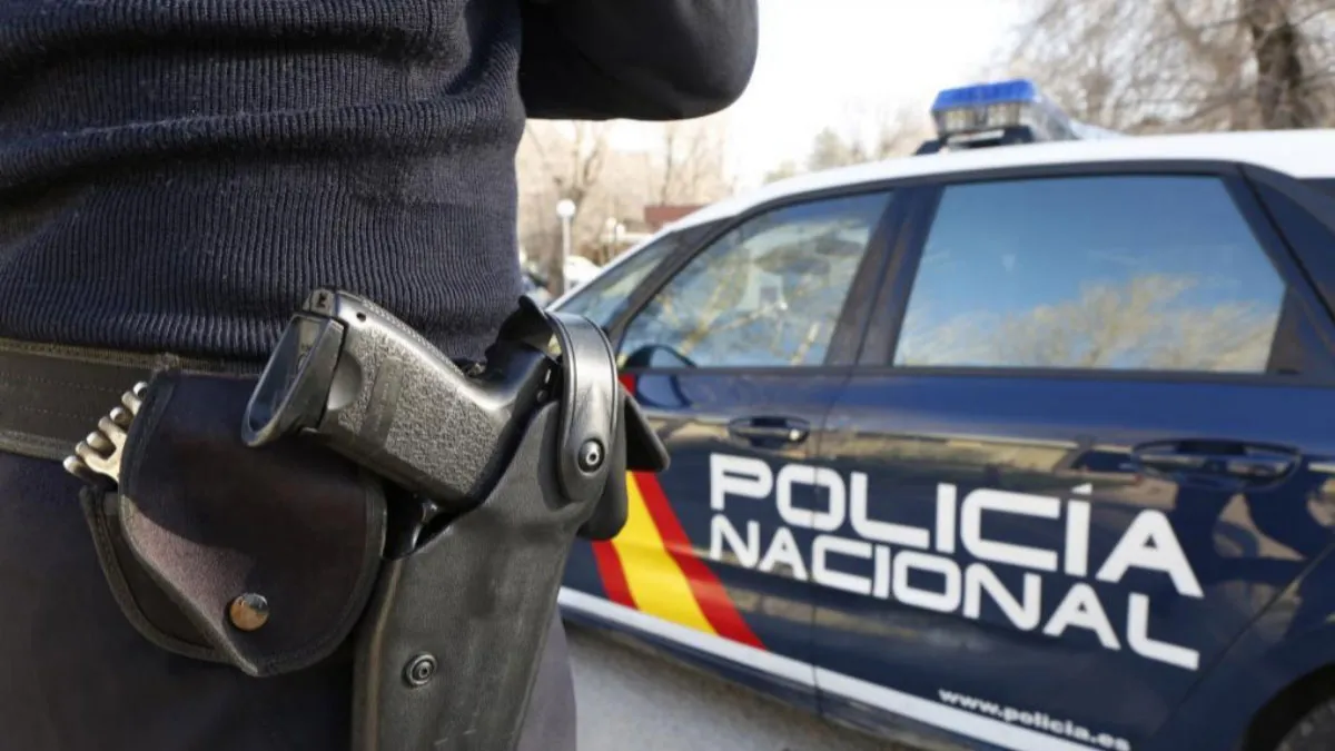 Cadera de un Policía Nacional con su arma reglamentaria y vehículo policial al fondo.