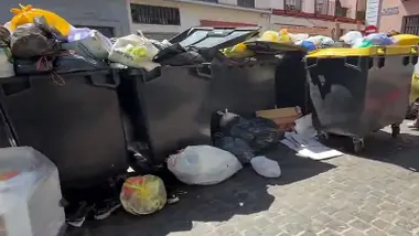 Contenedores grises con tapa amarilla llenos de basura, desbordados.
