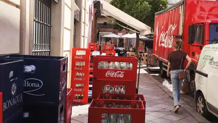 Camiones descargando cajas de bebidas en el centro. Son de Coca-Cola, destaca el rojo con letras blancas.